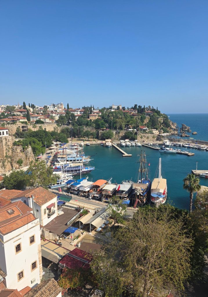 Elevated view over Antalya old town marina with tour boats and yachts anchored in deep blue water. Red roofed buildings and green cliffs surround the harbor while the Mediterranean Sea stretches beyond the breakwater.