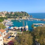 Panoramic view over Antalya old town marina with tour boats and yachts docked in turquoise water. Red roofed buildings and green cliffs surround the harbor as the Mediterranean Sea stretches beyond the breakwater.
