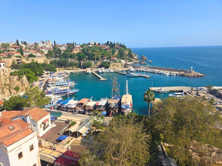 Panoramic view over Antalya old town marina with tour boats and yachts docked in turquoise water. Red roofed buildings and green cliffs surround the harbor as the Mediterranean Sea stretches beyond the breakwater.