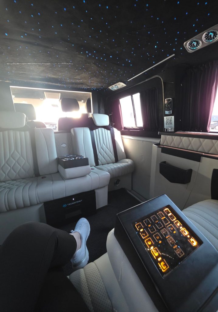 Luxury transfer van interior in Antalya with white leather seats. A blue star effect ceiling. Purple curtains. And an illuminated control panel beside a sign reading "ITS TRANSFER" and "WI FI".