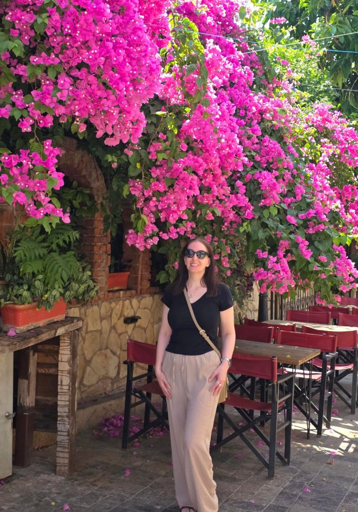 Me wearing sunglasses stands beneath vivid pink bougainvillea beside an outdoor restaurant in Antalya. Red chairs and wooden tables sit under the flowers against a rustic stone wall.