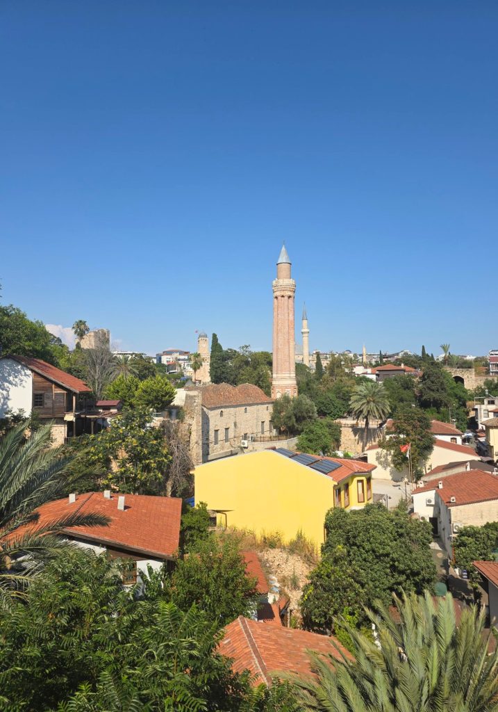 Elevated view across Antalya old town with red tiled roofs. Green trees. Historic stone buildings. And several minarets rising beneath a clear blue sky.