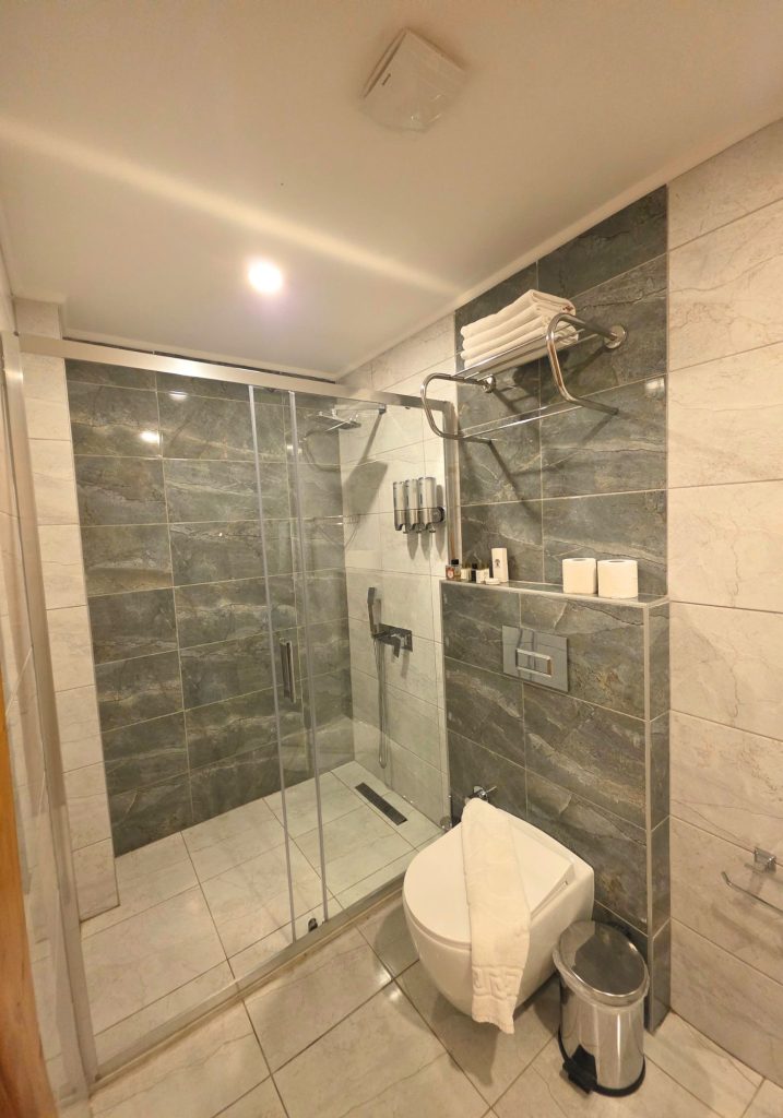 Modern hotel bathroom with gray stone effect tiles. A glass enclosed walk in shower. Folded towels. Toiletries. And a wall mounted toilet.