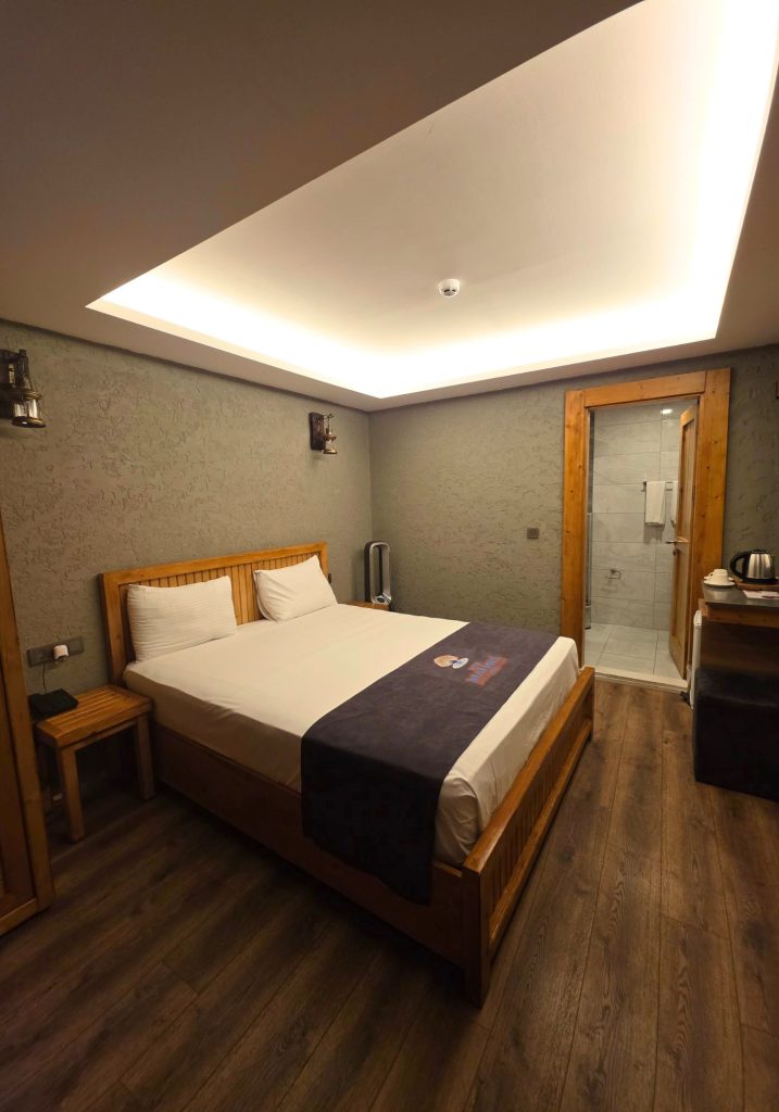 Warmly lit hotel room with a wooden double bed. Textured gray walls. Wood flooring. A kettle station. And an open doorway leading to a tiled bathroom.
