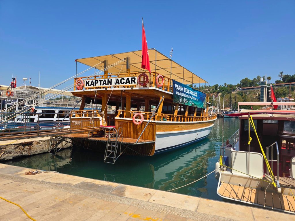 Wooden tour boat named "KAPTAN ACAR" docked in Antalya marina beneath a bright blue sky. A banner reads "YAPAY RESİF PROJESİ" and "Antalya'da Deniz Hep Temiz" while Turkish flags and life rings decorate the upper deck.