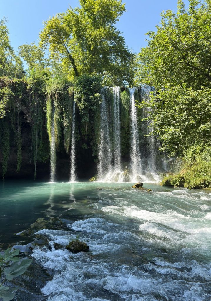 A wide waterfall in Antalya pours over a vine covered cliff into a turquoise pool. Fast moving water rushes around mossy rocks beneath tall leafy trees.