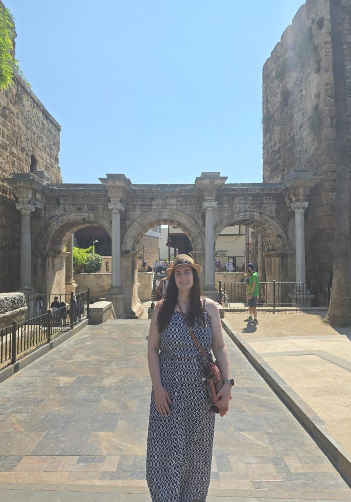 Me in a patterned maxi dress and sun hat stands before Hadrians Gate in Antalya. Three carved Roman arches and tall stone walls frame the busy pedestrian entrance.