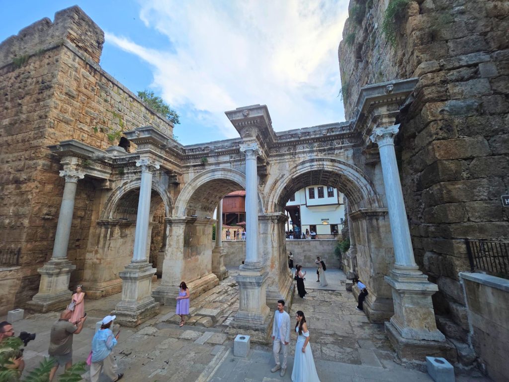 Wide view of Hadrians Gate in Antalya with three ornate Roman arches and tall marble columns between ancient stone walls. Visitors explore the worn passage while a couple in wedding attire poses beneath the central arch.