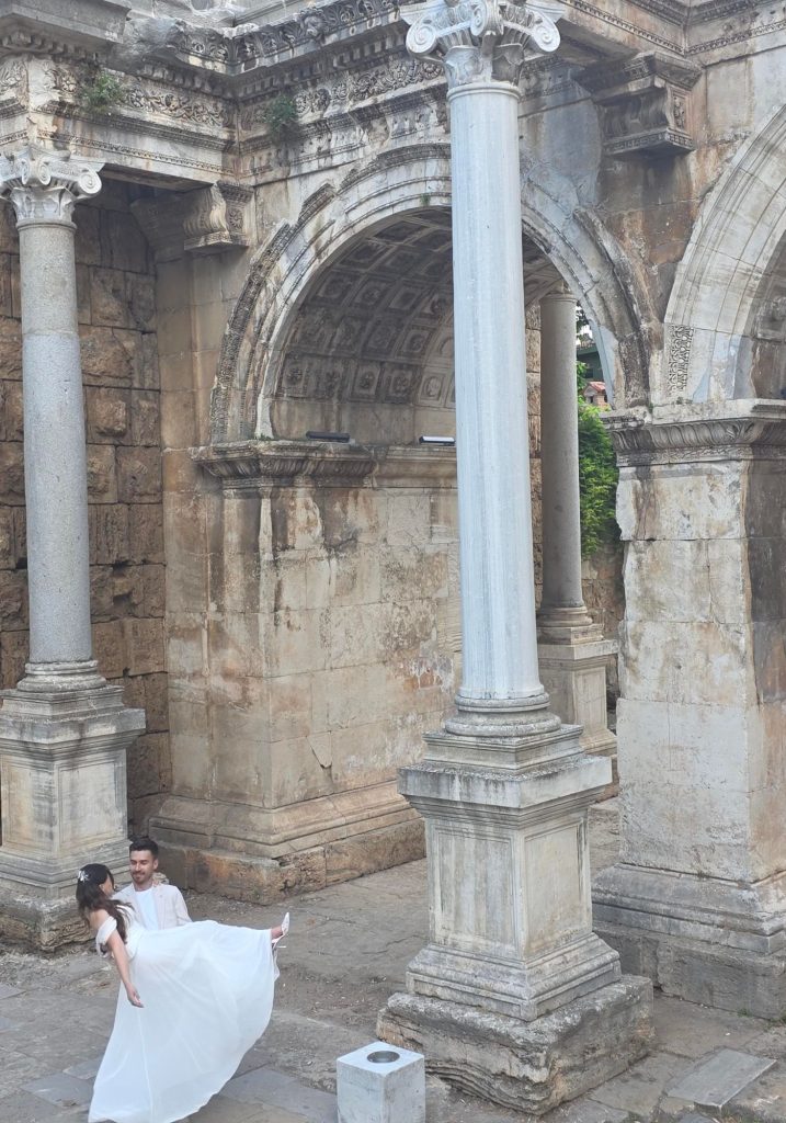 Ornate columns and carved stone arches at Hadrians Gate in Antalya. A groom carries a bride in a white wedding dress beneath the ancient Roman structure.