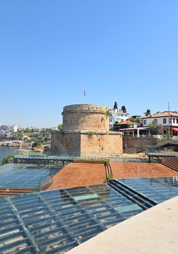 Hidirlik Tower in Antalya topped with a Turkish flag. Glass walkways and covered ruins fill the foreground with waterfront buildings and the sea behind it.