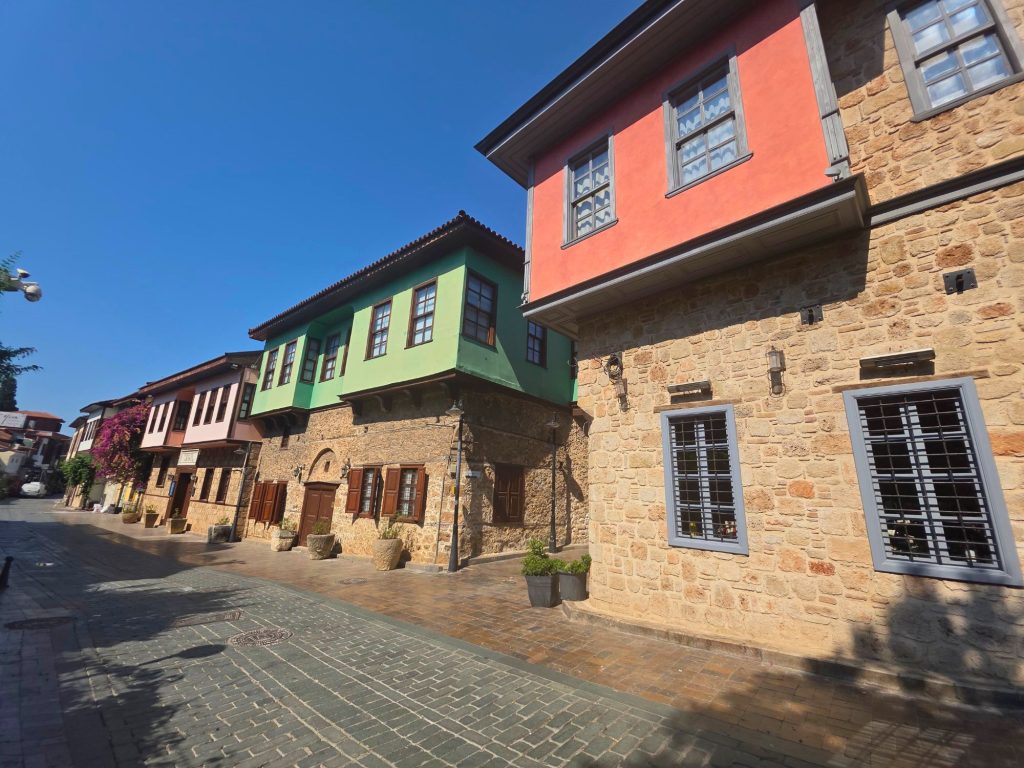 Sunlit cobblestone street in Antalya lined with restored Ottoman style houses. The buildings feature stone lower walls and colorful green red and pale pink upper floors beneath a clear blue sky.
