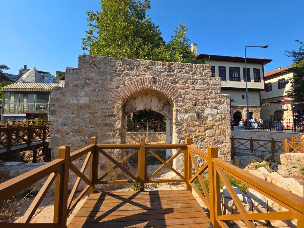 Ancient stone arch and excavated ruins in Antalya framed by wooden walkways and railings. Traditional buildings and leafy trees rise behind the archaeological site under a clear blue sky.
