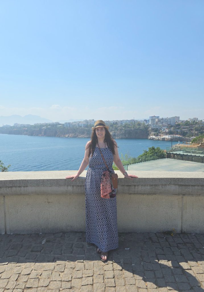 Me in a patterned maxi dress and sun hat poses at a stone overlook in Antalya. The Mediterranean Sea cliffs and city skyline stretch across the background under a clear blue sky.