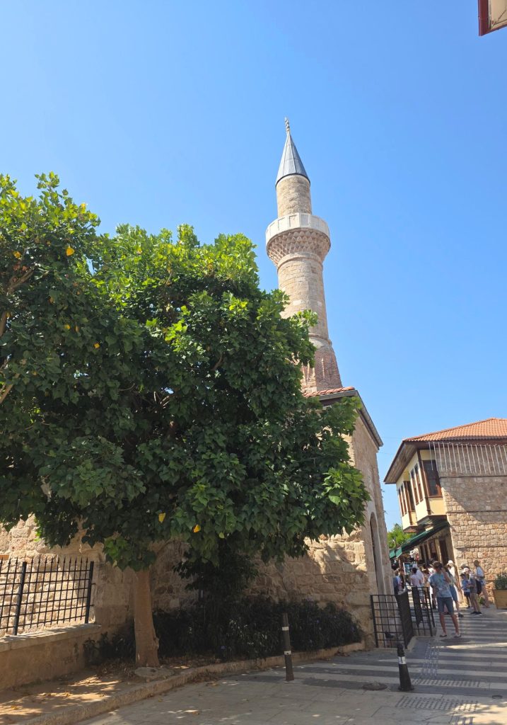 Slender stone mosque minaret rises above leafy trees and traditional buildings in Antalya. Visitors walk along the shaded street below on a bright sunny day.