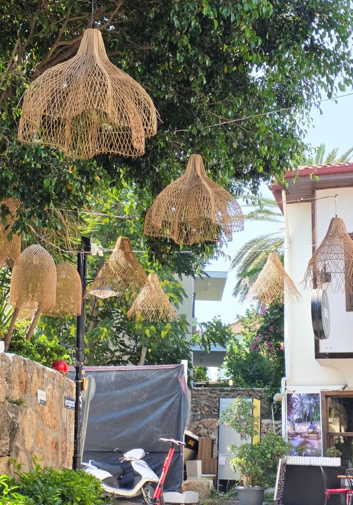 Woven rattan lampshades hang beneath leafy trees along a narrow lane in Antalya. A white scooter and red electric scooter sit beside stone walls and a white building.