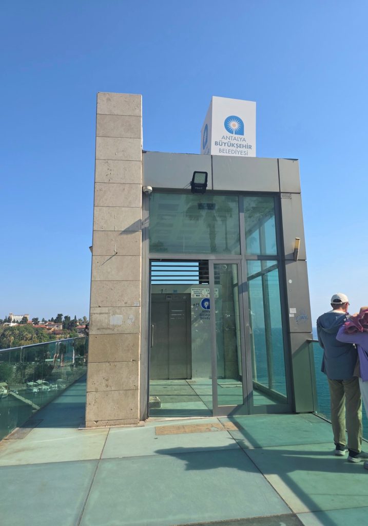 Glass elevator entrance on a coastal viewing platform in Antalya beneath a clear blue sky. The sign reads "ANTALYA BÜYÜKŞEHİR BELEDİYESİ" and two visitors stand beside the railing overlooking the sea.