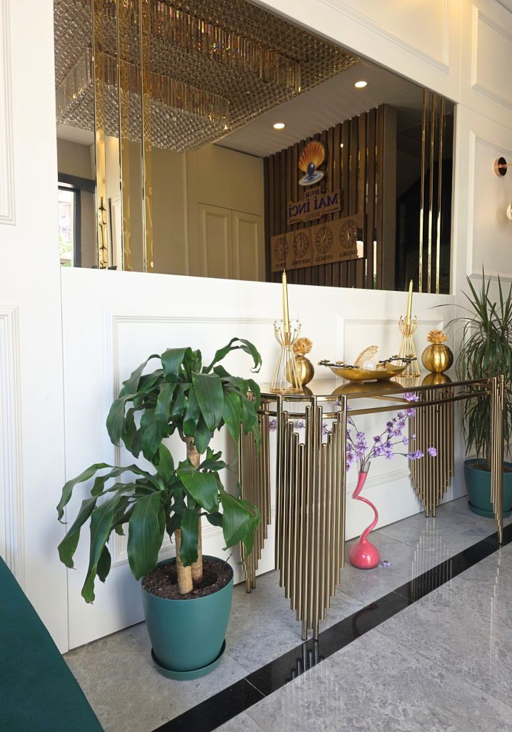 Decorative Antalya hotel lobby with gold furniture. Large green plants. Purple flowers. And a mirrored wall reflecting the sign "BER MALİNCİ HOTEL" with clocks labeled "LONDRA" "ANTALYA" "NEWYORK" and "TOKYO".