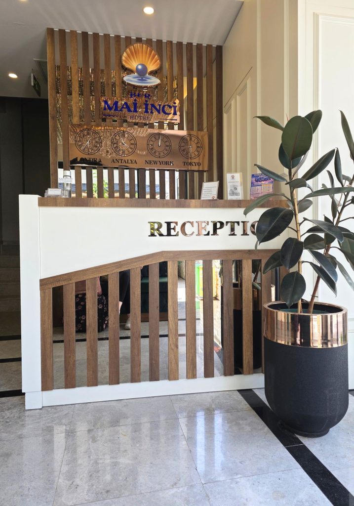 Bright reception area at an Antalya hotel with a white and wood front desk and a large potted rubber plant. Signs behind the desk read "BER MALİNCİ HOTEL" and "LONDRA" "ANTALYA" "NEWYORK" "TOKYO" while the desk displays "RECEPTION".