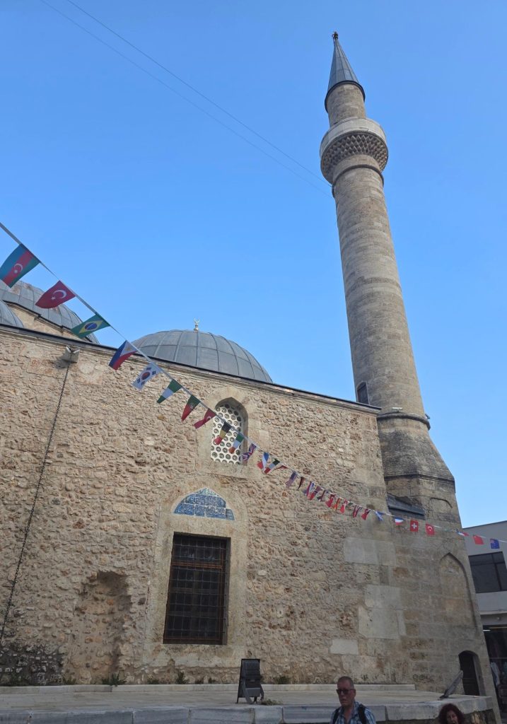 Slender stone mosque minaret rises above leafy trees and traditional buildings in Antalya. Visitors walk along the shaded street below on a bright sunny day.