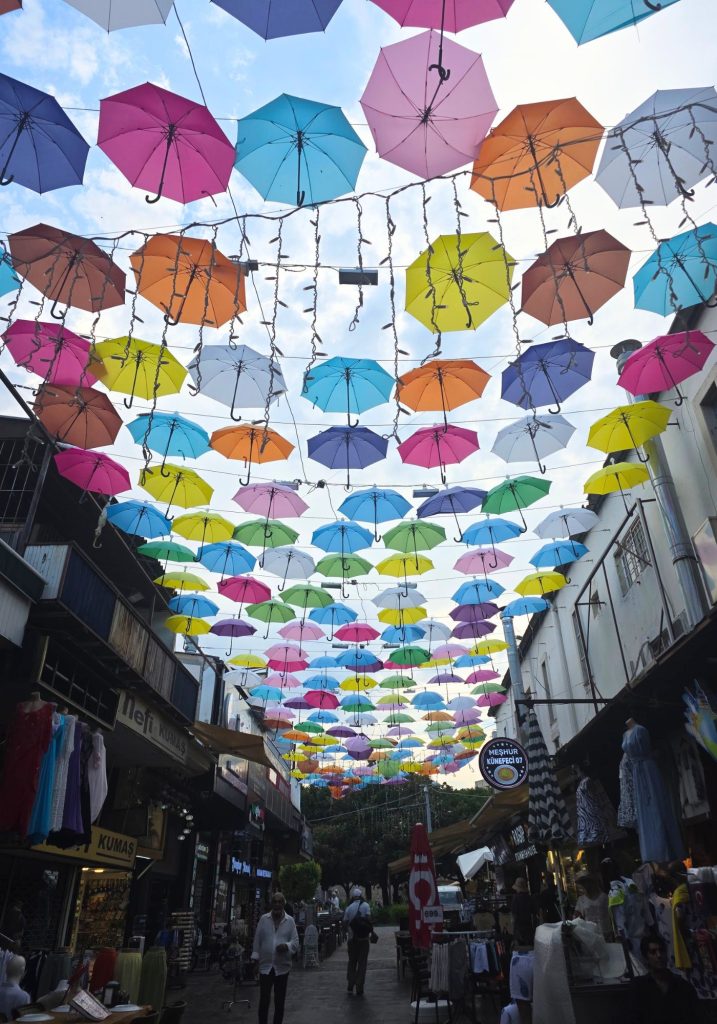 Colorful umbrellas and strings of lights form a canopy over a busy shopping street in Antalya. Clothing shops restaurants and pedestrians line the shaded pedestrian lane.