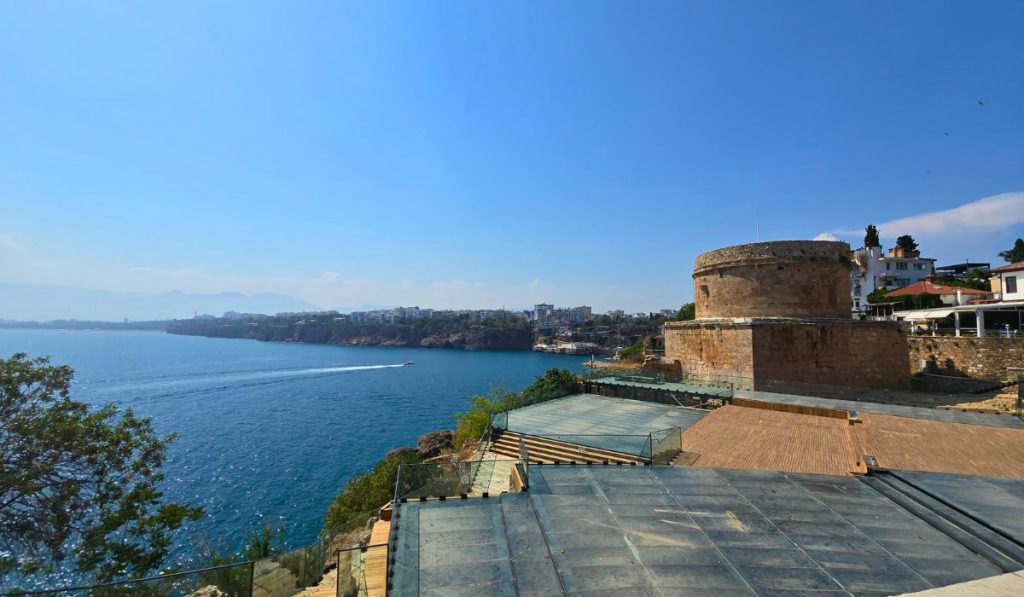 anoramic view across the blue Mediterranean coastline in Antalya with a speedboat leaving a white wake. A round stone tower and waterfront buildings sit above glass covered archaeological ruins.