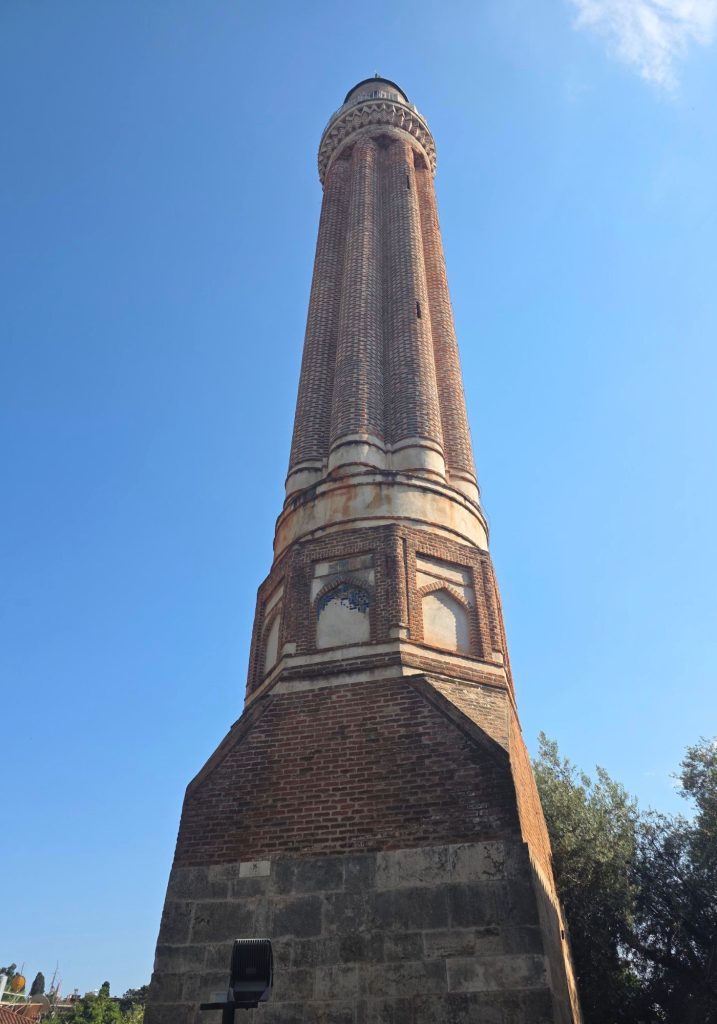 Stone mosque and tall minaret photographed from below in Antalya. Strings of international flags cross the courtyard beneath the domed roof and clear blue sky.
Image: uploaded image 4.jpg
Alt text: Tall fluted brick minaret in Antalya viewed from its broad stone base. Decorative brickwork and a carved upper balcony stand out against the cloudless blue sky.