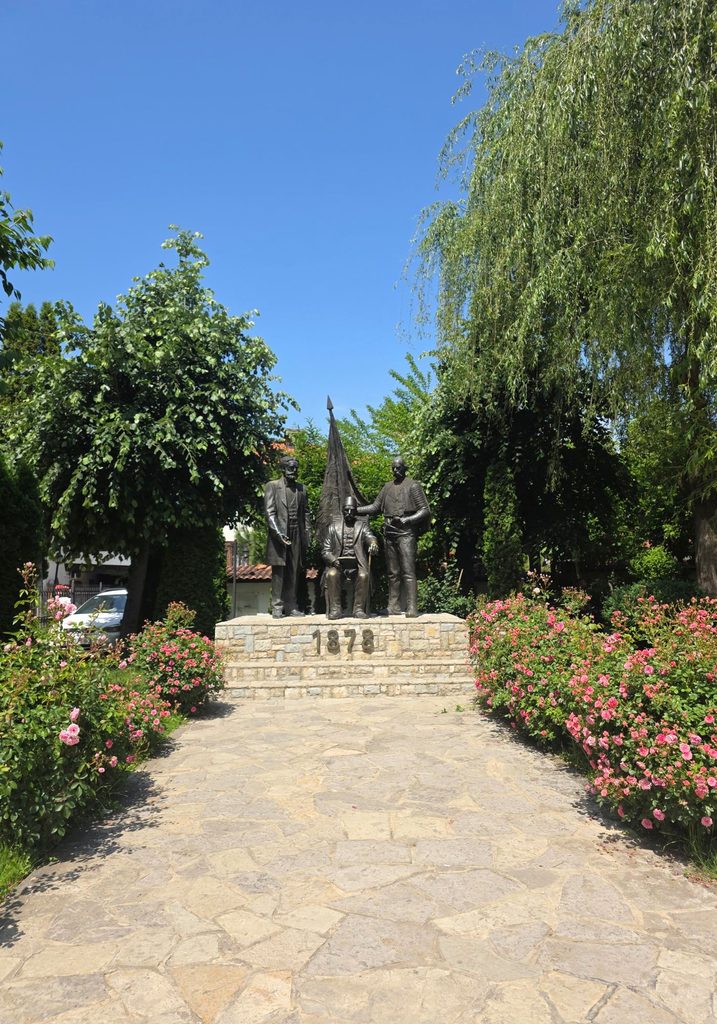 Bronze figures on a stone monument at the Albanian League of Prizren complex, surrounded by flowers and trees.