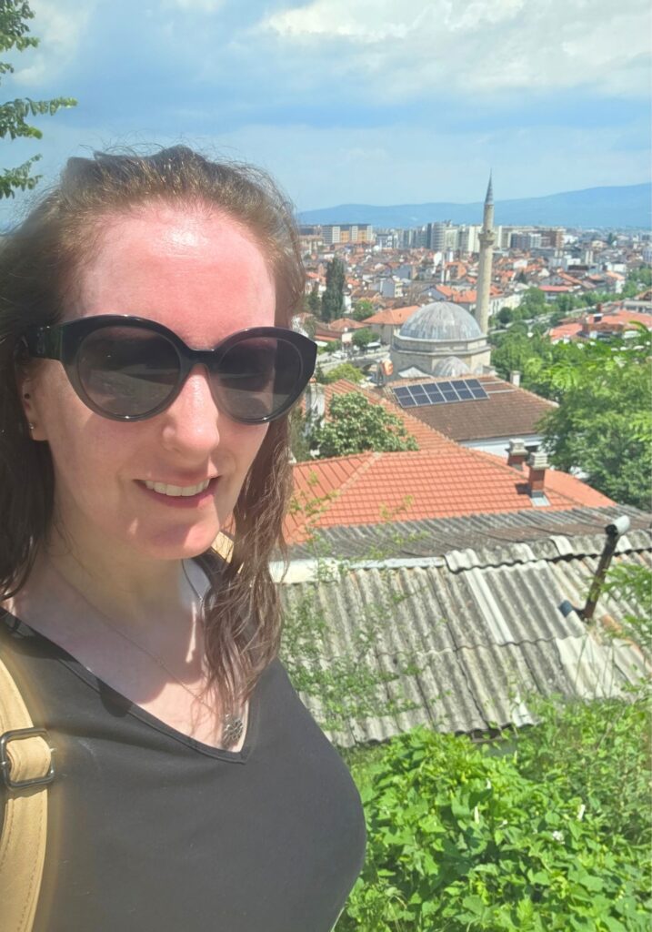Me wearing sunglasses at a hillside viewpoint in Prizren, with red rooftops, a domed mosque and mountains behind me.
