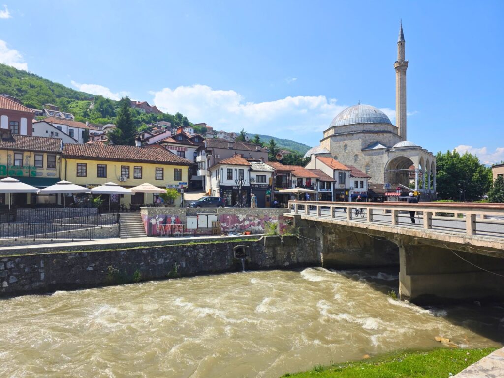 River flowing beneath a bridge in central Prizren, with riverside buildings and a domed mosque beyond.