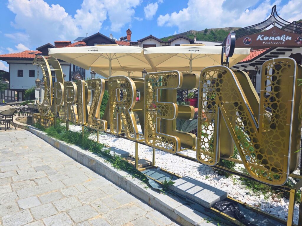 Large gold PRIZREN letters beside a paved walkway, with cafés and red-roofed buildings behind them.
