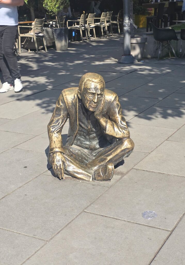 Bronze sculpture of a man sitting cross-legged on the pavement in Pristina, beside outdoor café tables.