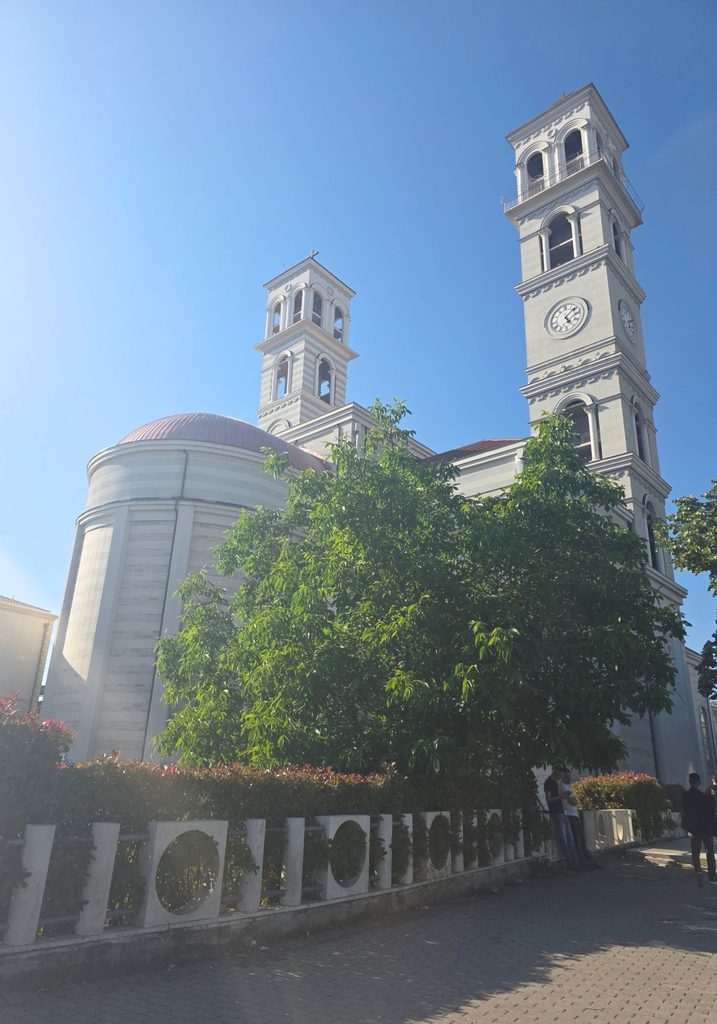 Side view of Mother Teresa Cathedral in Pristina, showing its rounded apse and two bell towers behind trees.