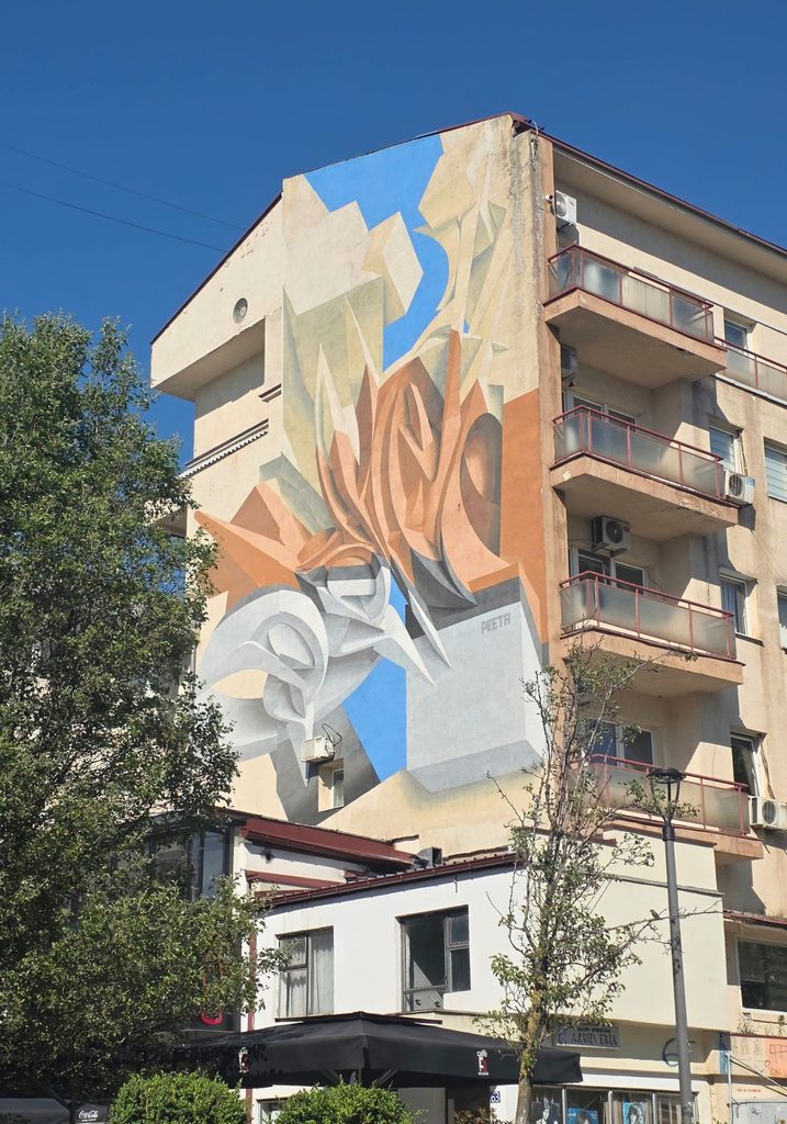 Abstract mural by Peeta covering the side of an apartment building in Pristina, with flowing geometric shapes.
